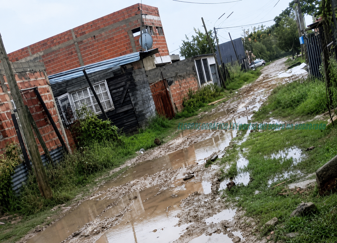 Barrios de Berisso, crítico relevamiento de Armendi revela abandono en calles 22 Fotografía documental de una calle de tierra anegada en un barrio de Berisso tras la lluvia, con pozos de agua visibles y viviendas de fondo. "barrios de Berisso calles anegadas reclamo vecinal".
